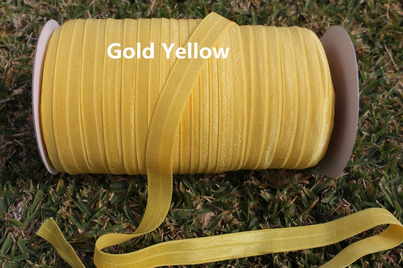 May include: A spool of gold yellow elastic fold over elastic, also known as FOE, is shown on a white cardboard spool. The elastic is wound tightly and is ready to be used for sewing projects.