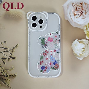 May include: A white phone case with a clear back and a floral design. The design features a cartoon bear with flowers and leaves. The case has the text "Pretty flowers" printed around the edge.