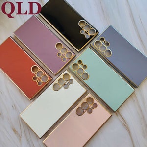May include: A set of eight phone cases in various colors, including black, pink, red, white, light blue, and purple. Each case has a gold trim and a cutout for the camera lens.