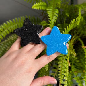 May include: Two star-shaped objects with a smooth, glossy finish. One star is black with fine silver glitter particles throughout, and the other star is a vibrant blue with fine silver glitter particles throughout the surface.
