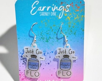May include: Clear acrylic earrings with silver hooks. Each earring features a cartoon image of a Propofol bottle with the text "Just Go with the FLO". The background is a gradient of blue, pink, and purple.