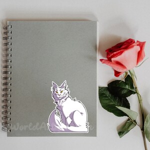 May include: A white cat with yellow eyes sits on a gray notebook with a spiral binding. A pink rose with green leaves is on the right side of the image.