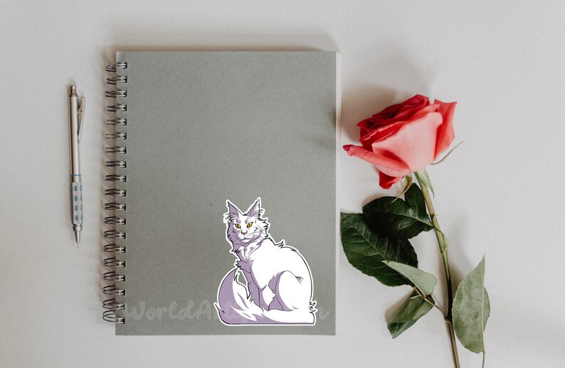 May include: A white cat with yellow eyes sits on a gray notebook with a spiral binding. A pink rose with green leaves is on the right side of the image.