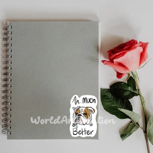 May include: A white sticker with a cartoon bulldog wearing a collar and the text "Ah... Much Better". The sticker is on a gray notebook with a spiral binding. A pink rose is on the right side of the image.