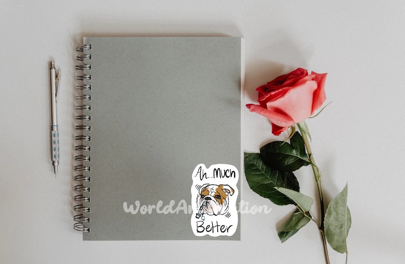 May include: A white sticker with a cartoon bulldog wearing a collar and the text "Ah... Much Better". The sticker is on a gray notebook with a spiral binding. A pink rose is on the right side of the image.