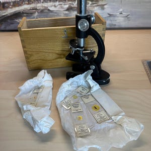 May include: A black metal microscope with a silver eyepiece and adjustment knobs, positioned in front of a light-colored wooden storage box. Several glass microscope slides with white labels are resting on white tissue paper in the foreground.
