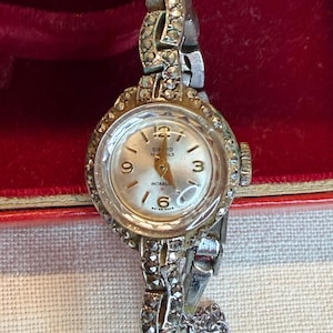 Könnte beinhalten: Eine silberfarbene Armbanduhr mit rundem Zifferblatt in Weiß, goldfarbenen Zeigern und Ziffern bei 12, 3, 6 und 9. Die Lünette und das Armband sind mit kleinen, dunkelgrauen Steinen besetzt. Das Zifferblatt zeigt den Text Sinos, 17 Jewels, Incabloc und Swiss Made.