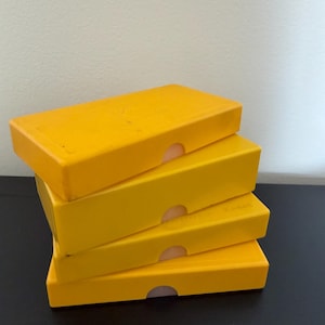 May include: A stack of four rectangular yellow plastic storage cases. Each case features a smooth, solid yellow exterior with a small semi-circular cutout on the front edge, designed to facilitate opening the lid of each individual container.