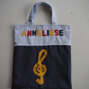 May include: A blue and white gingham tote bag with the name "Anneliese" embroidered in colorful letters. The bag has a black and white polka dot fabric lining with a yellow treble clef.