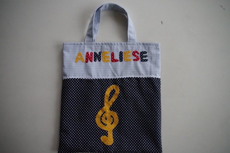 May include: A blue and white gingham tote bag with the name "Anneliese" embroidered in colorful letters. The bag has a black and white polka dot fabric lining with a yellow treble clef.