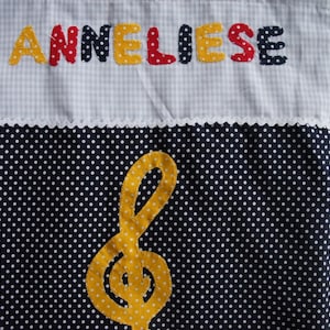 May include: A white and blue gingham fabric bag with the name "ANNELIESE" embroidered in yellow, red, and blue letters. The bag has a white lace trim and a black fabric with white polka dots on the bottom. A yellow felt musical note is centered on the polka dot fabric.