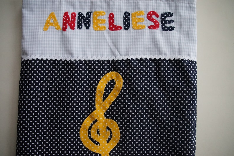May include: A white and blue gingham fabric bag with the name "ANNELIESE" embroidered in yellow, red, and blue letters. The bag has a white lace trim and a black fabric with white polka dots on the bottom. A yellow felt musical note is centered on the polka dot fabric.