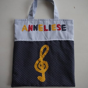 May include: A blue and white gingham tote bag with a black and white polka dot bottom. The bag has a yellow treble clef and the name "Anneliese" spelled out in colorful letters.