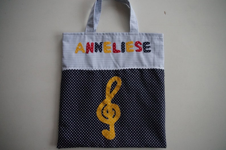 May include: A blue and white gingham tote bag with a black and white polka dot bottom. The bag has a yellow treble clef and the name "Anneliese" spelled out in colorful letters.