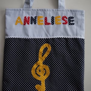 May include: A blue and white gingham tote bag with the name "Anneliese" spelled out in colorful letters. The bag has a white trim and a black and white polka dot bottom with a yellow treble clef.