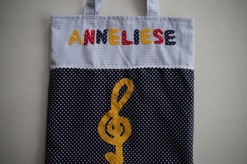 May include: A blue and white gingham tote bag with the name "Anneliese" spelled out in colorful letters. The bag has a white trim and a black and white polka dot bottom with a yellow treble clef.