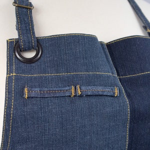 Può includere: Un grembiule in denim blu con cuciture gialle e un anello di metallo nero sul cinturino. Il grembiule ha una sola tasca sul davanti.