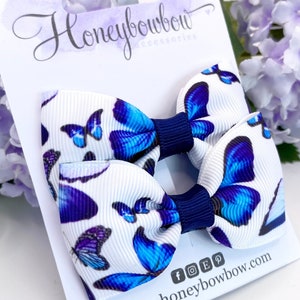 Puede incluir: Dos pinzas para el pelo de mariposa azul y blanca con centros azul marino. Las pinzas están en una tarjeta blanca con el texto "Honeybowbow Accessories" y "www.honeybowbow.com".