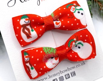 May include: Two red bow hair clips with a white polka dot pattern and snowman design. The clips are displayed on a white card with the text "Honeybowbow accessories". The background includes Christmas decorations.