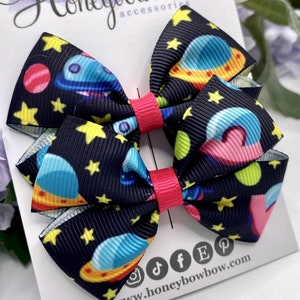 May include: Two black and white hair bows with a space-themed print. The bows feature spaceships, planets, and stars. The bows are tied with pink ribbon.