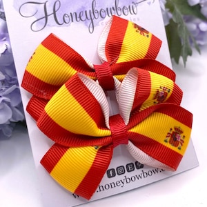 May include: Two small red, yellow, and white hair bows with the Spanish flag and coat of arms printed on them. The bows are on a white card with the text "Honeybowbow" printed on it.