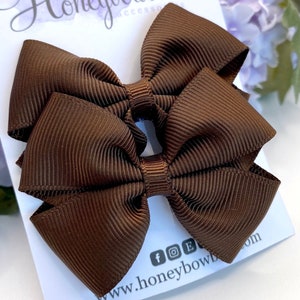 May include: Two brown hair bows made of ribbed fabric. The bows are neatly arranged on a white card with the text "Honeybow Accessories" and social media icons. The bows are a rich, solid brown color.