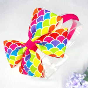 May include: A colourful rainbow hair bow with a scale pattern, attached to a bright pink headband. The bow features a spectrum of colours including red, orange, yellow, green, blue, and purple. The bow is made of a ribbon material.