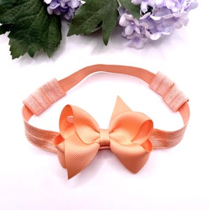 May include: A peach-colored baby headband with a large bow. The bow is made of a ribbed fabric and is centered on a peach-colored elastic band. The headband is designed for infants and toddlers.