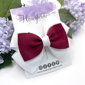 May include: A white and claret hair bow with a layered design. The bow features a white base with a claret centre section. The bow is attached to a white card with the text "Honeybowbow access" and social media icons.
