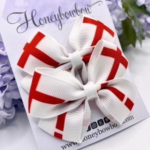 May include: Two white and red hair bows with a cross pattern. The bows are attached to a white card with the text "Honeybowbow Accessories" and "www.honeybowbow.com".