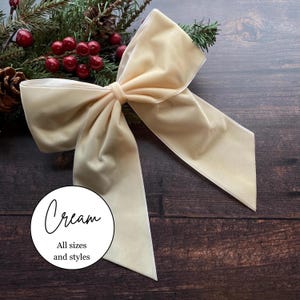 May include: A cream-coloured velvet bow with long, flowing tails. A circular white label is positioned in the lower left corner with the text "Cream" in a script font above the printed text "All sizes and styles" in a simple sans-serif font.