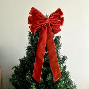 May include: A large, festive red velvet bow with long, flowing tails, atop a green Christmas tree. The bow has a layered design with a contrasting gold trim, creating a luxurious holiday decoration. The bow is a vibrant accent for seasonal decor.