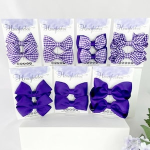 May include: Six display cards featuring pairs of hair bows in various purple shades. The top row shows purple and white gingham bows, while the bottom row displays solid purple bows. Each card is branded with the text Honeybowbow at the top.