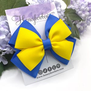May include: A blue and yellow hair bow with a layered design. The bow has a yellow centre and blue outer layers. The bow is on a white card with the text "Honeybowbow accessor" and social media icons.