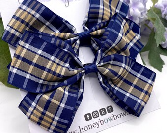 May include: Two navy blue plaid hair bows with tan and white accents. The bows are on a white card with the text "Honeybow accessories" and social media icons. A sprig of purple flowers is in the background.