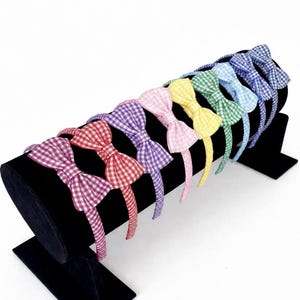 May include: A selection of gingham bow headbands in a variety of colours, including pink, red, purple, yellow, green, and blue. The headbands are displayed on a black stand. The bows are a classic, charming accessory.