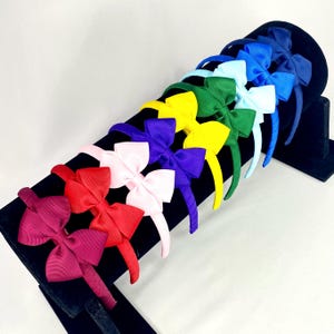 May include: A selection of headbands with large bows in a variety of colours. The headbands are displayed in a rainbow sequence, including purple, pink, red, yellow, green, and blue. The bows are made of a ribbed material.