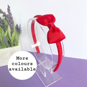 May include: A red headband with a bow. The headband is displayed on a clear stand. The bow is made of a ribbed fabric. The text "More colours available" is in a white circle.