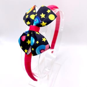 Puede incluir: Una diadema rosa con un lazo negro con un estampado espacial colorido con estrellas, planetas y cohetes.
