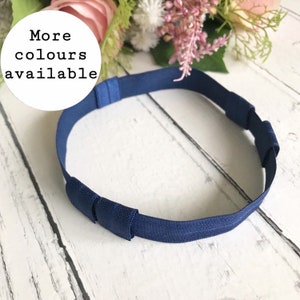May include: A navy blue headband with adjustable elastic straps. The text "More colours available" is in a white circle in the upper left corner. The headband is on a white wooden surface.