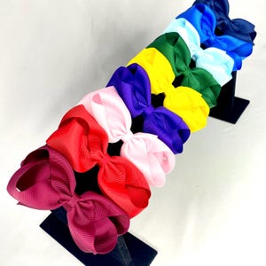 May include: A selection of colourful hair bows in shades of red, pink, purple, yellow, green, and blue. The bows are presented on black stands against a white backdrop, highlighting their vibrant colours and textures.
