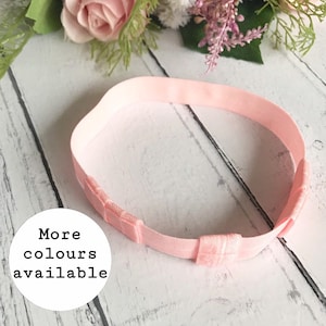Puede incluir: Una diadema ajustable de color rosa claro. La diadema está hecha de un material suave y elástico y presenta un diseño sencillo. El texto "More colours available" está en un círculo blanco. El fondo es una superficie de madera clara con flores rosas.