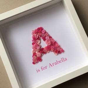 May include: A framed artwork featuring the letter "A" crafted from various shades of pink buttons. The text "is for Arabella" is printed in pink below the letter. The artwork is set in a white shadow box frame.