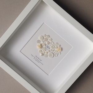 May include: A white frame with a white background and a heart-shaped design made of white buttons. The number 30 is in the center of the heart. The text "Ann & Carl! Happy Pearl Wedding Anniversary 2nd August 2018" is below the heart.