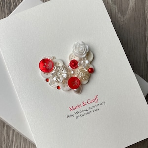 May include: A white card with a red and white heart made of buttons, beads, and a white flower. The text on the card reads "Marie & Geoff Ruby Wedding Anniversary 6th October 2024".