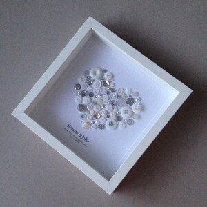 May include: A white frame with a white background and a heart shape made of white and silver buttons. The text "Silvana & John" is written below the heart.