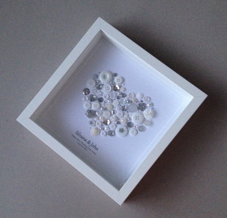 May include: A white frame with a white background and a heart shape made of white and silver buttons. The text "Silvana & John" is written below the heart.