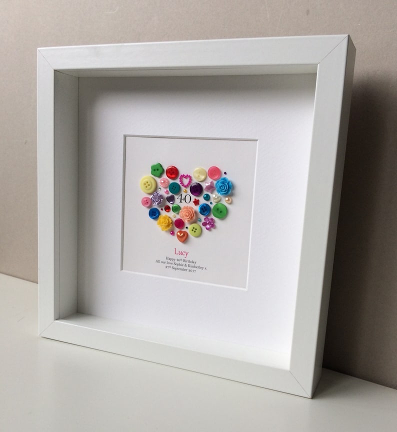 Puede incluir: Un marco blanco con un paspart&uacute; blanco que rodea un coraz&oacute;n hecho de botones coloridos. El coraz&oacute;n est&aacute; decorado con un n&uacute;mero 40 rosa y el texto "Lucy Happy 40th Birthday All our love Sophie & Kimberley 27th September 2017".