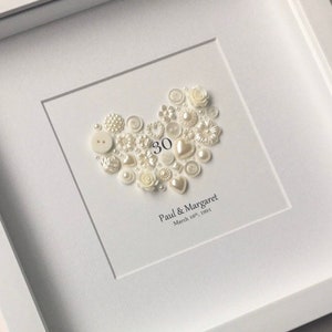 May include: A white framed picture with a heart made of white buttons and pearls. The number 30 is in the center of the heart. The text below the heart reads "Paul & Margaret March 18th, 1991".