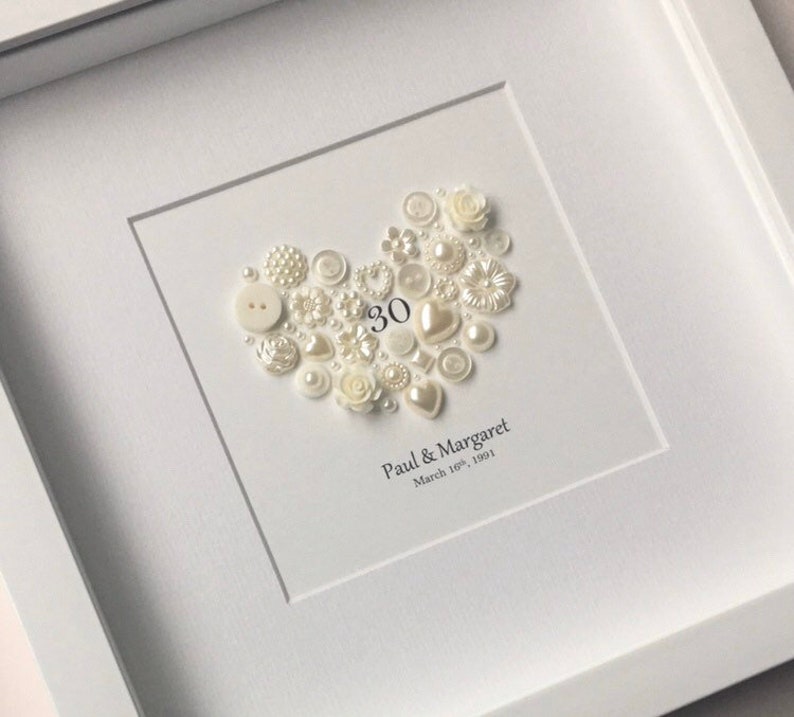 May include: A white framed picture with a heart made of white buttons and pearls. The number 30 is in the center of the heart. The text below the heart reads "Paul & Margaret March 18th, 1991".